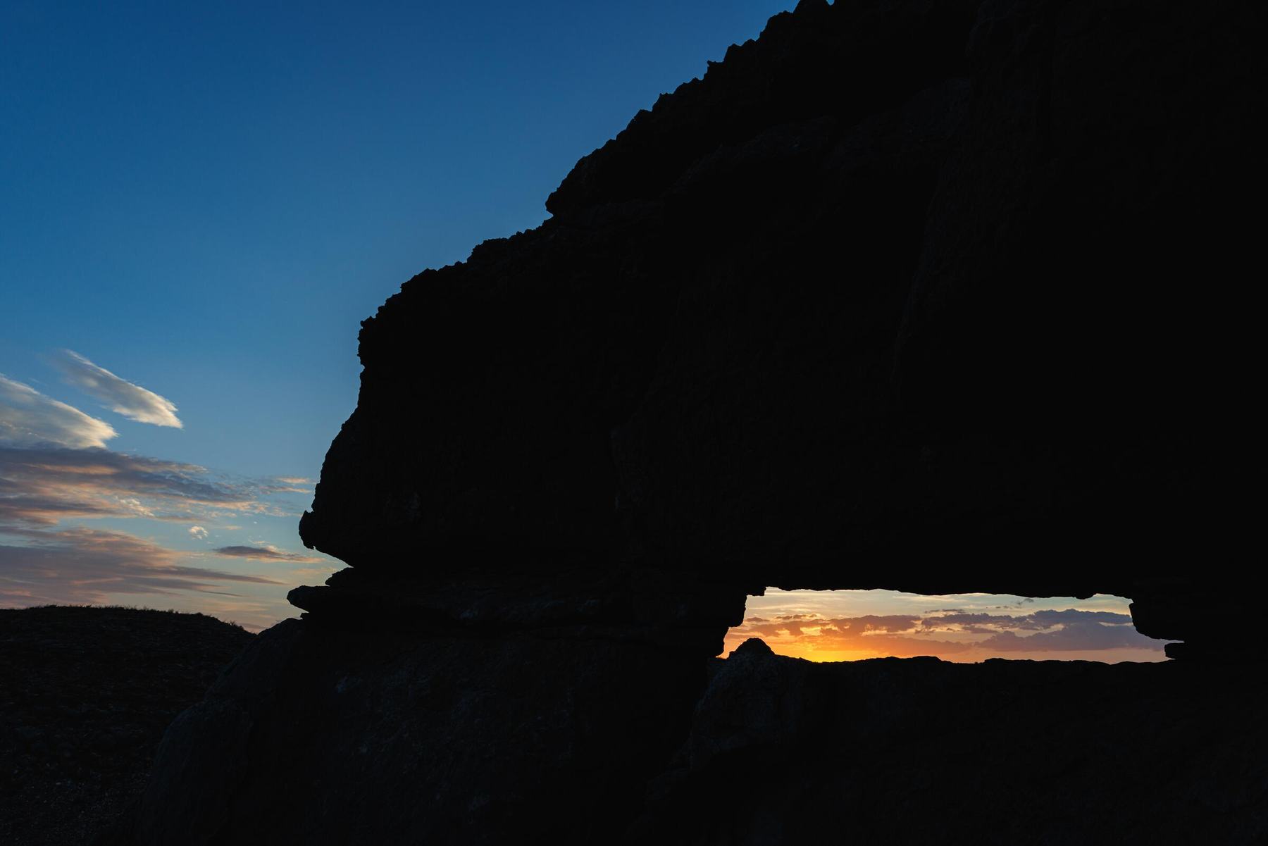 A dark rock ledge against a blue evening sky, with sunset glowing through a window in the rock